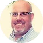 Portrait of a smiling bald man with glasses and a white beard, wearing a light striped shirt.