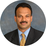 Studio headshot of a man in a dark suit, light blue shirt, and gold patterned tie against a gray backdrop