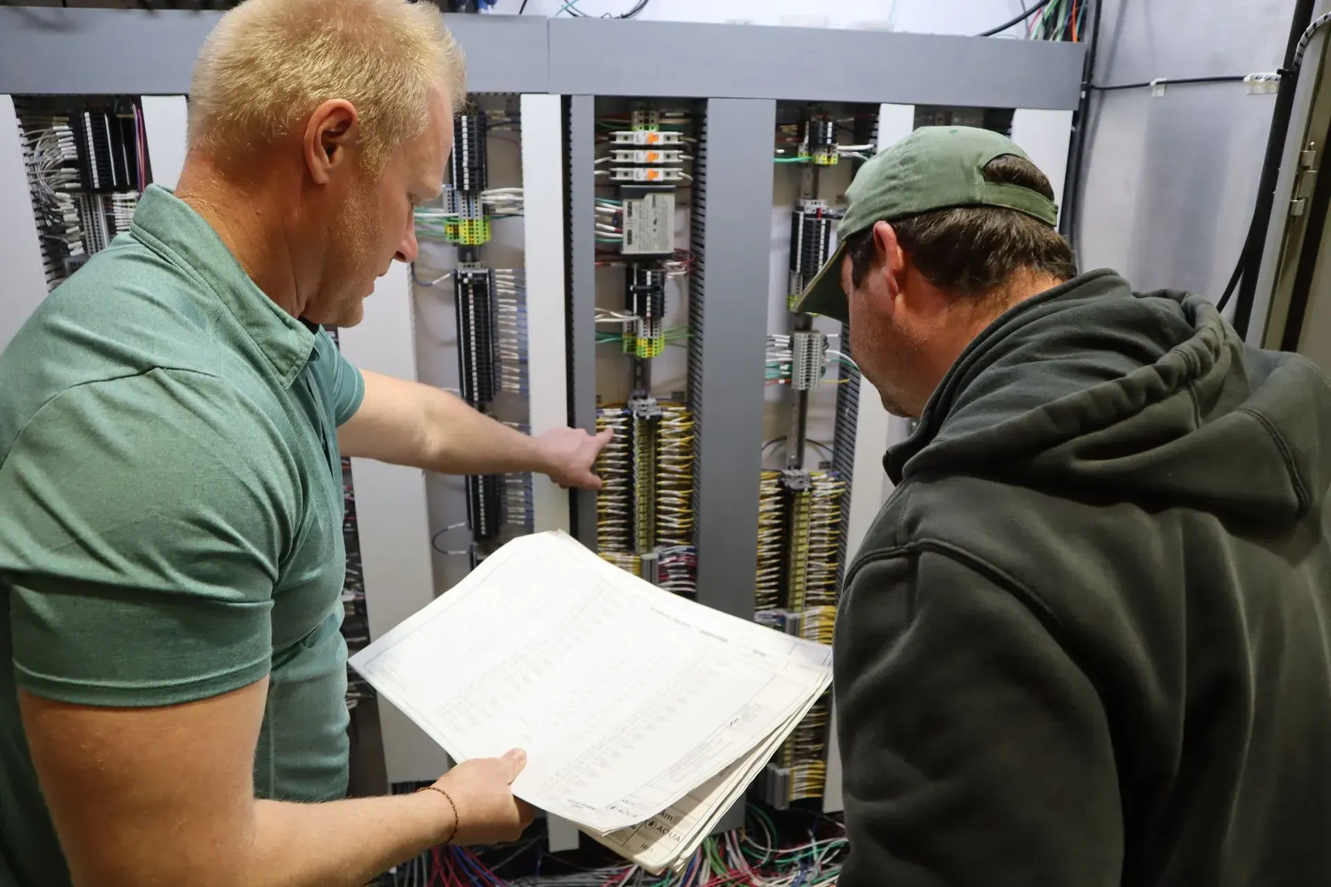 Two men are looking at a piece of paper in front of a electrical panel.