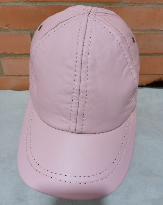 Gorra de béisbol rosa claro con costuras cosidas, mostrada de frente contra una pared de ladrillos.