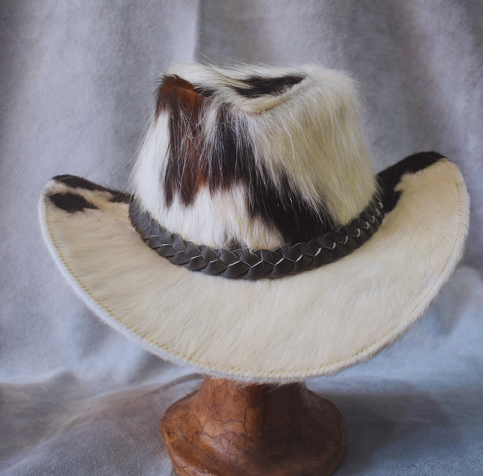 Sombrero de vaquero con pelo blanco, banda trenzada negra y copa de retazos marrones y negros sobre un soporte.