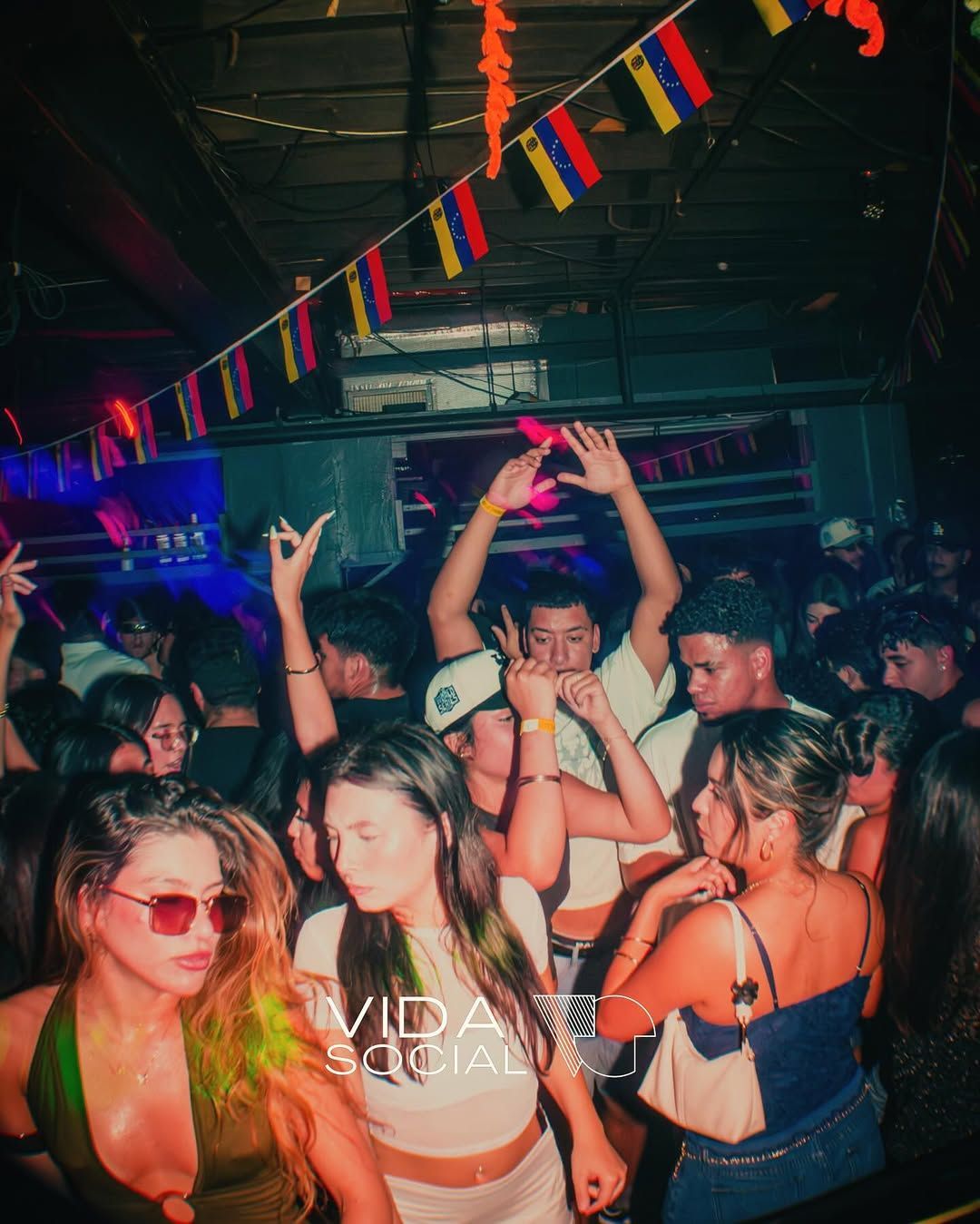 Crowded nightclub with people dancing under colorful hanging flags and neon lights.