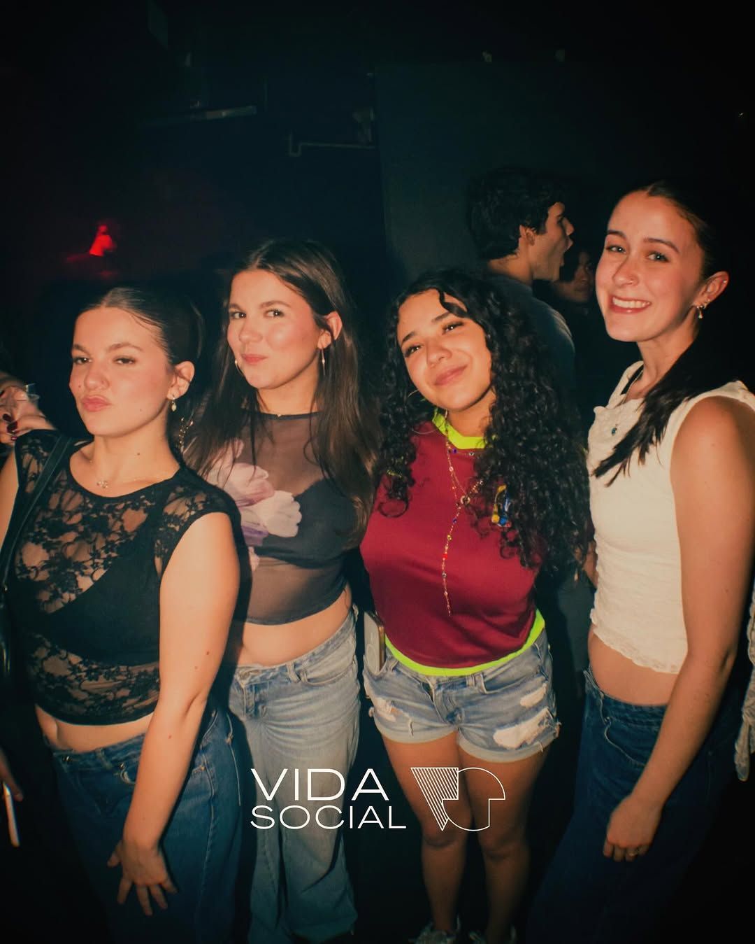 Four friends posing at a dark nightclub, smiling in colorful tops and jeans.