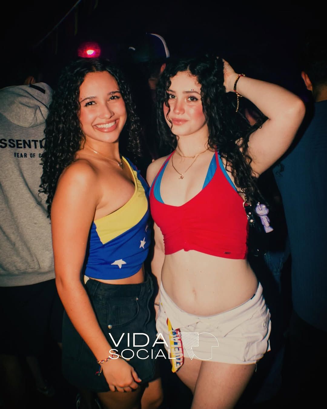 Two women posing at a nightclub, one in a blue-and-yellow top and one in a red top with beige shorts.