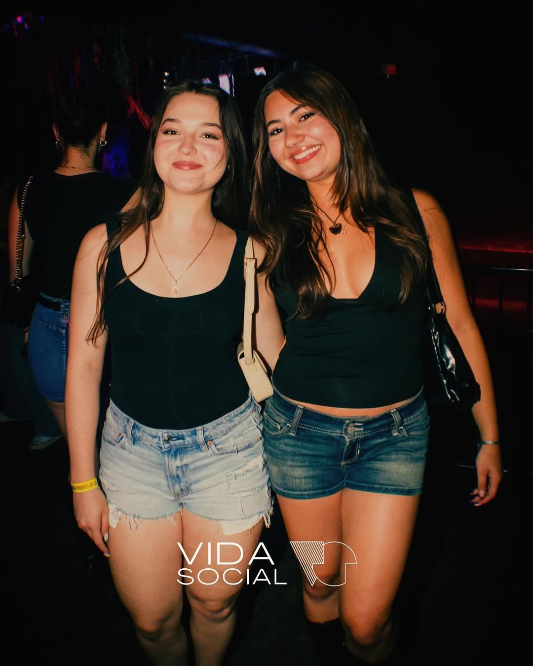 Two friends in black tops and denim shorts posing and smiling in a dim nightclub setting