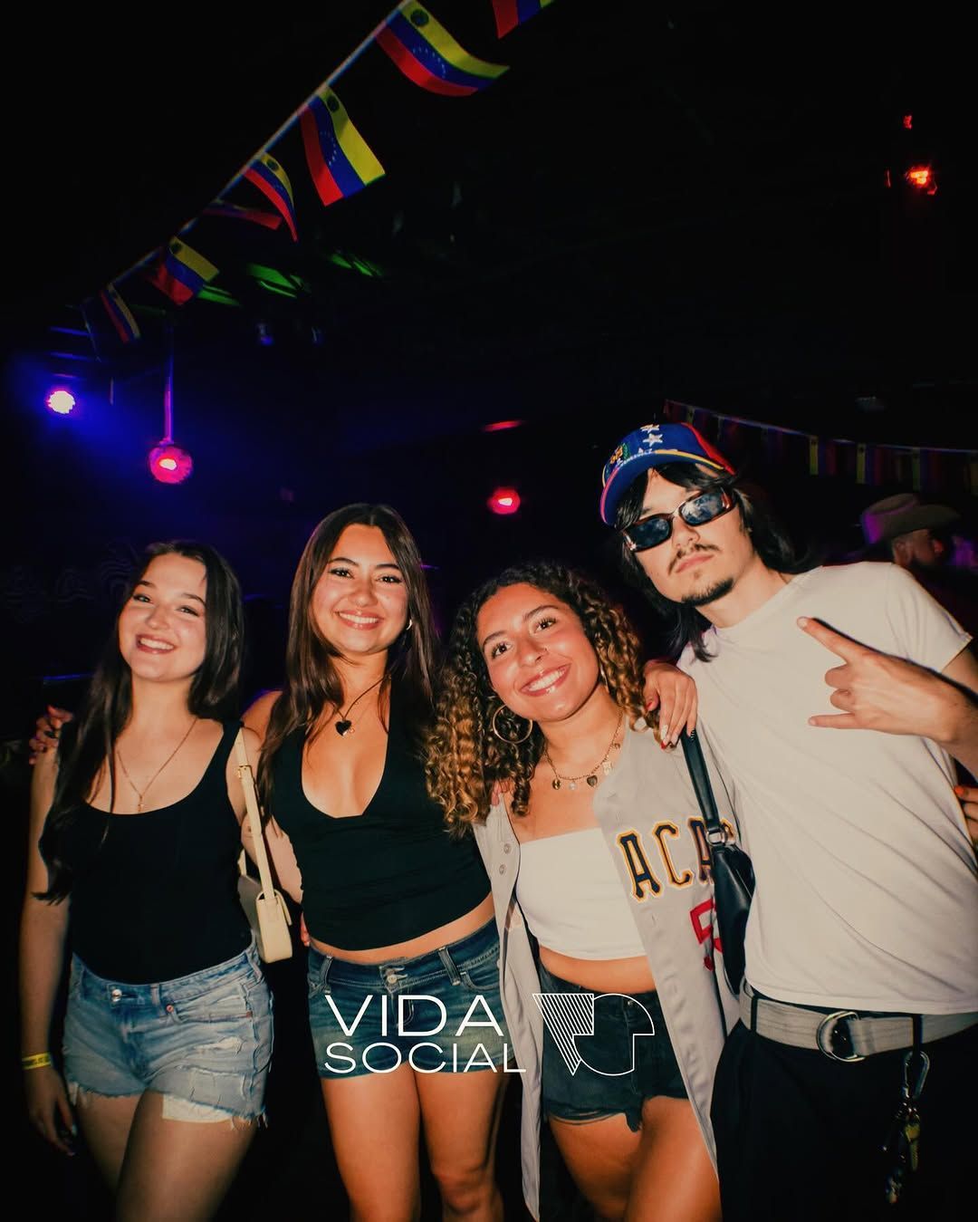Four friends posing at a dark party, wearing casual clothes and sunglasses under colorful lights.