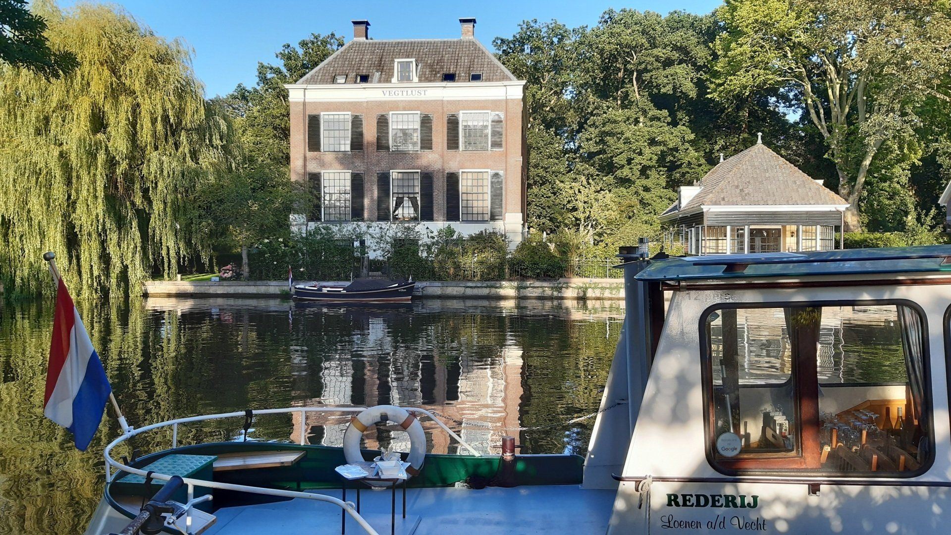 Boot  met uitzicht op een historisch huis, wilgenbomen en een Nederlandse vlag.