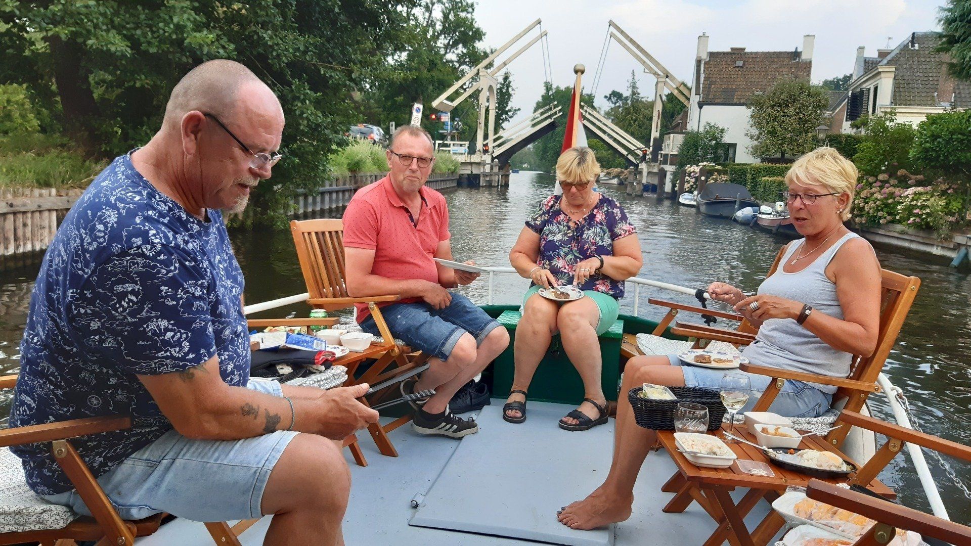 Vier mensen zitten op een boot en eten wat. Op de achtergrond zijn een brug en gebouwen te zien.