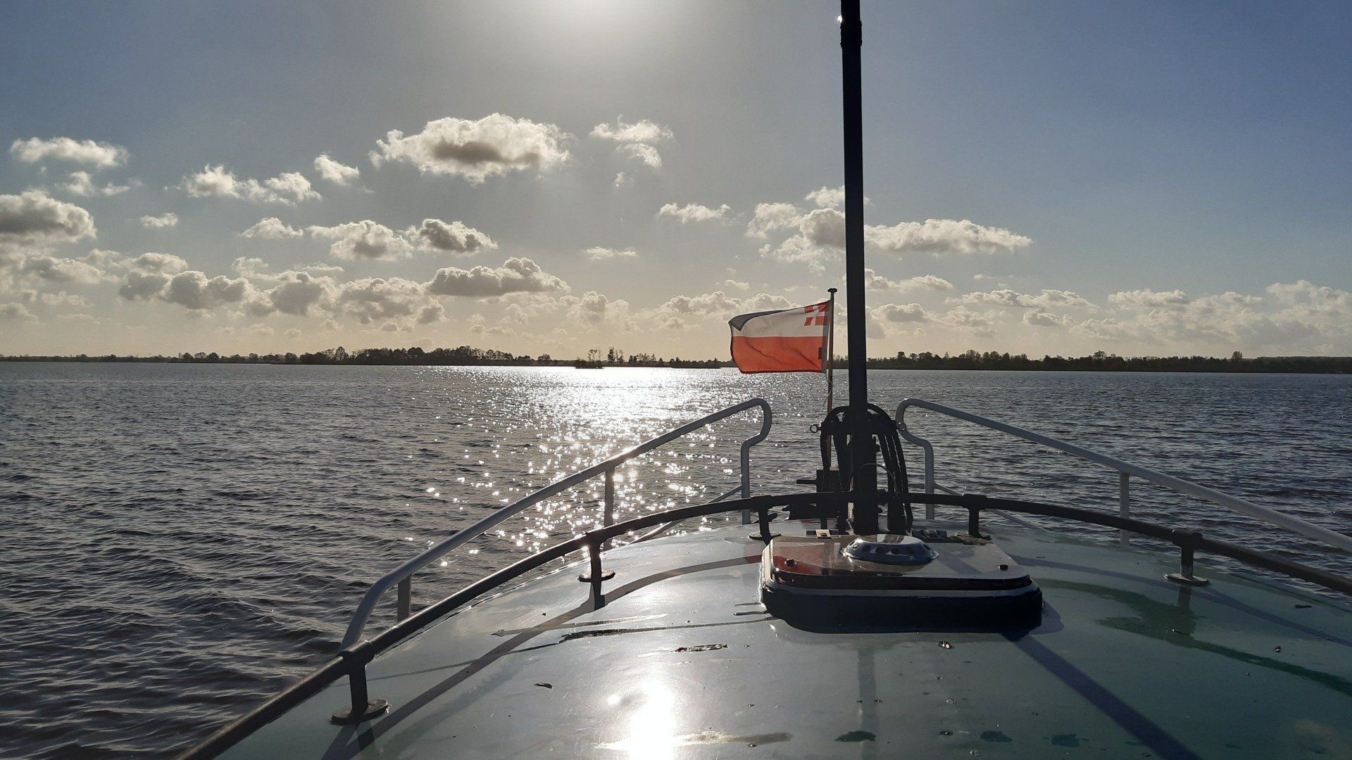 Een boot vaart onder een stralende zon richting een verre kustlijn, met de Poolse vlag in de mast.