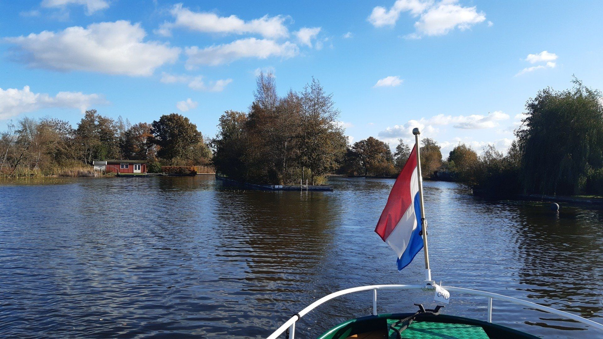 Boot op een kanaal met Nederlandse vlag; bomen en kleine gebouwen aan de oever; blauwe lucht.