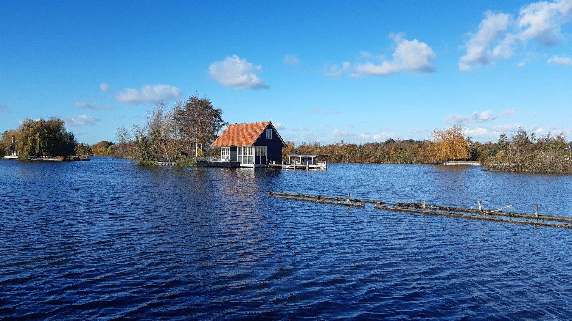 Een klein huisje met een oranje dak staat op het water, omringd door bomen onder een blauwe hemel.
