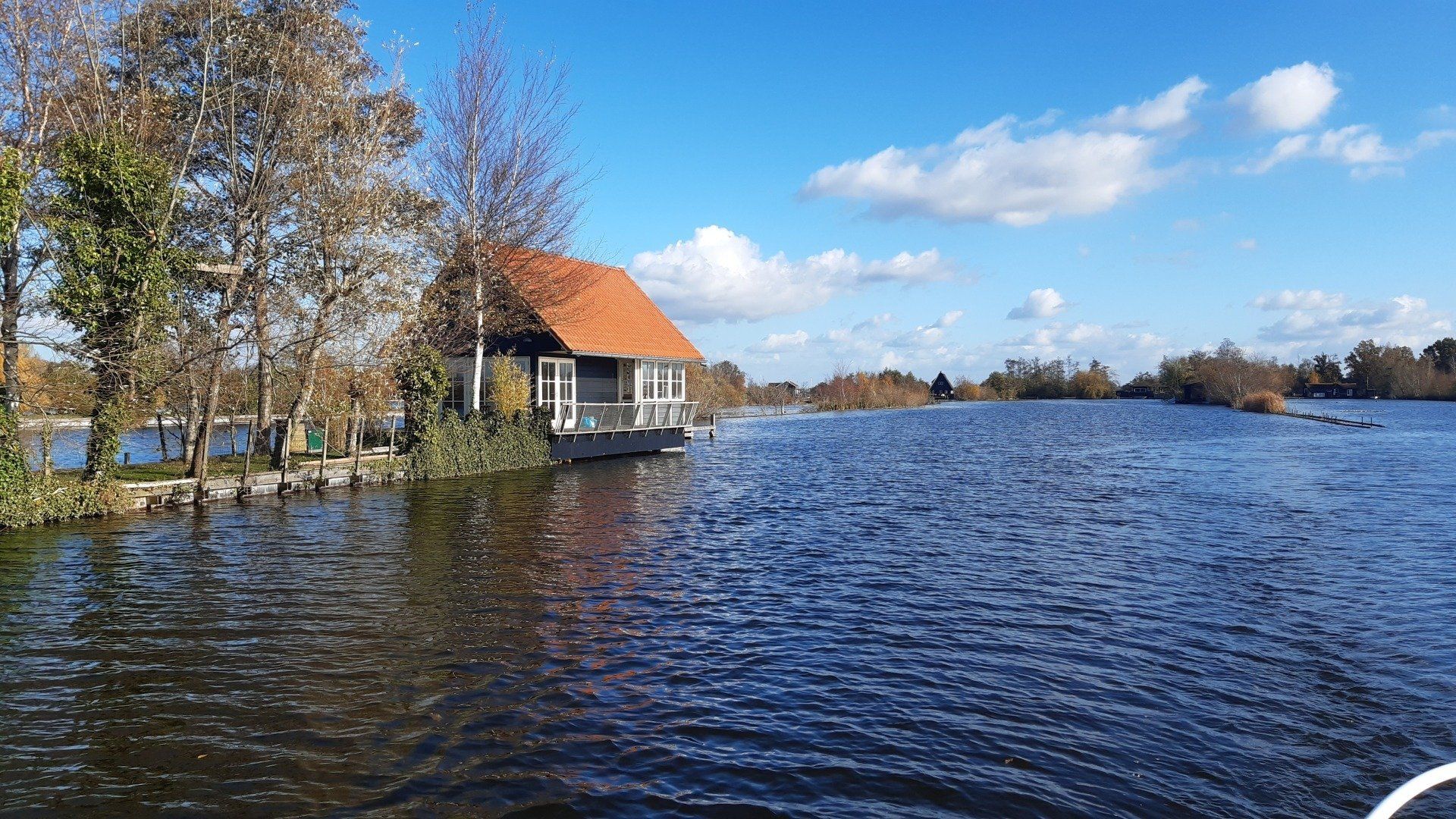Een klein huisje met een oranje dak staat in een waterrijk landschap onder een blauwe hemel met pluizige wolken.