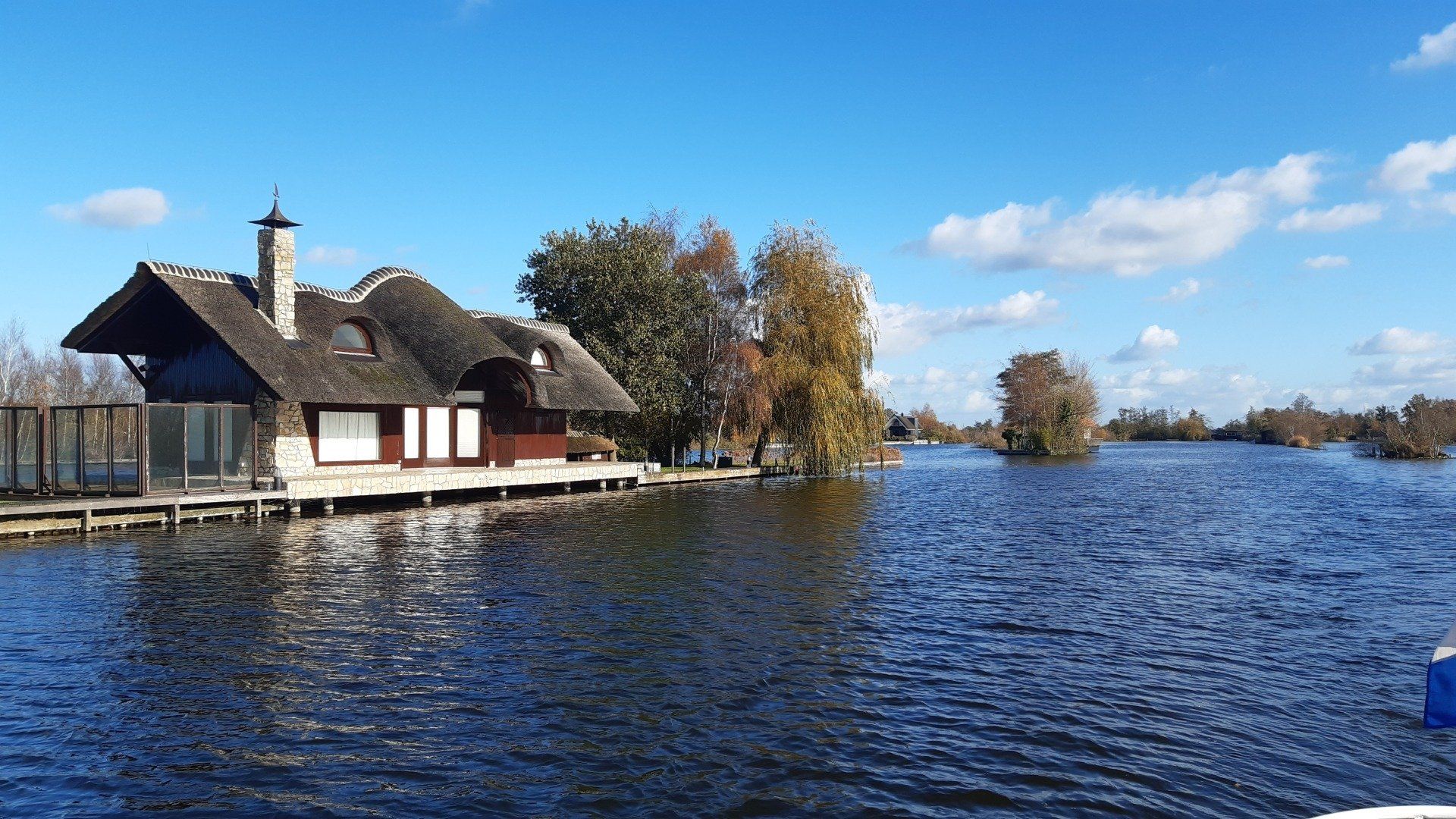 Een huisje aan het meer met een rieten dak en schoorsteen. Het huis is omgeven door water onder een blauwe hemel met wolken.