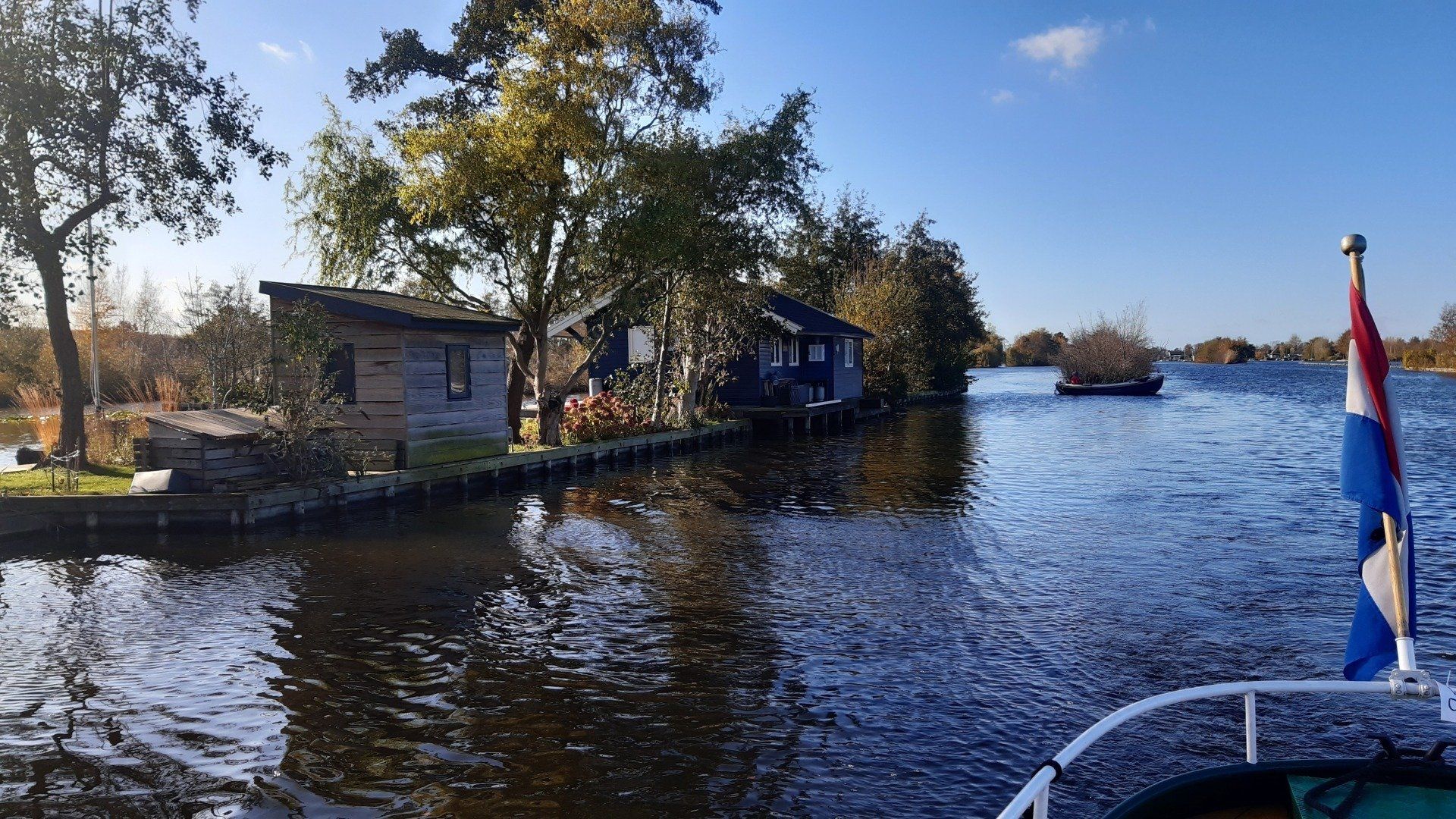 Kanaalscène met huizen en een boot, Nederlandse vlag op de boot, blauwe lucht.