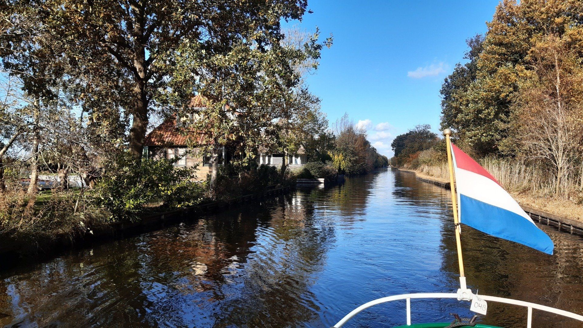 Een boot vaart over een kanaal dat omzoomd is met bomen en een huis. Een Nederlandse vlag wappert aan de boot.