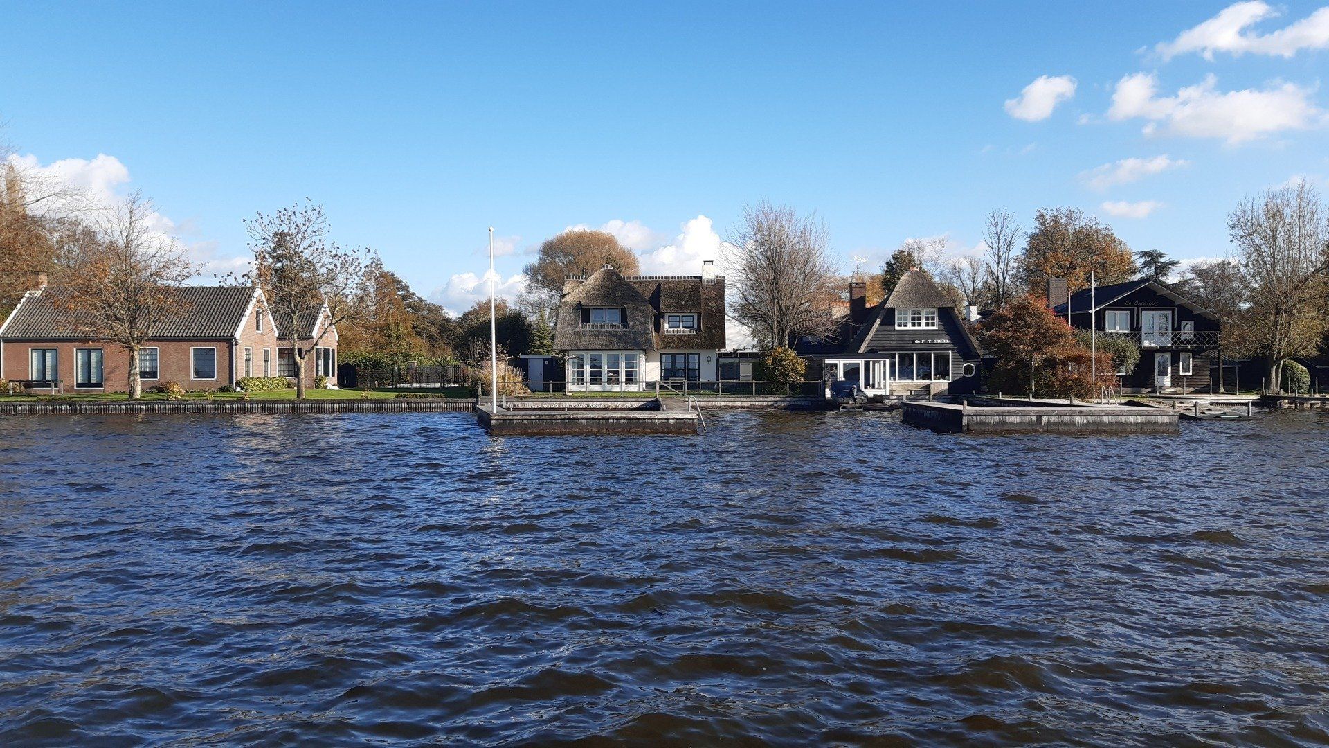 Huizen aan een meer onder een blauwe hemel, met bomen op de achtergrond. Het kalme water weerspiegelt het tafereel.