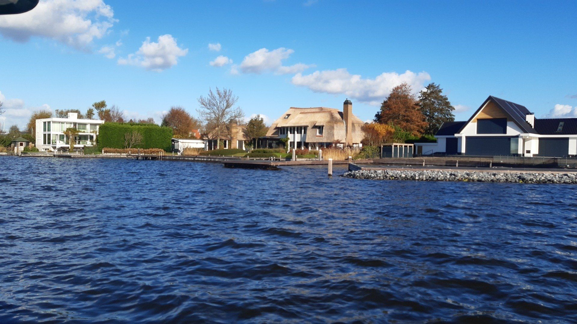 Huizen aan het meer onder een blauwe hemel, met kabbelend water op de voorgrond en herfstbladeren.