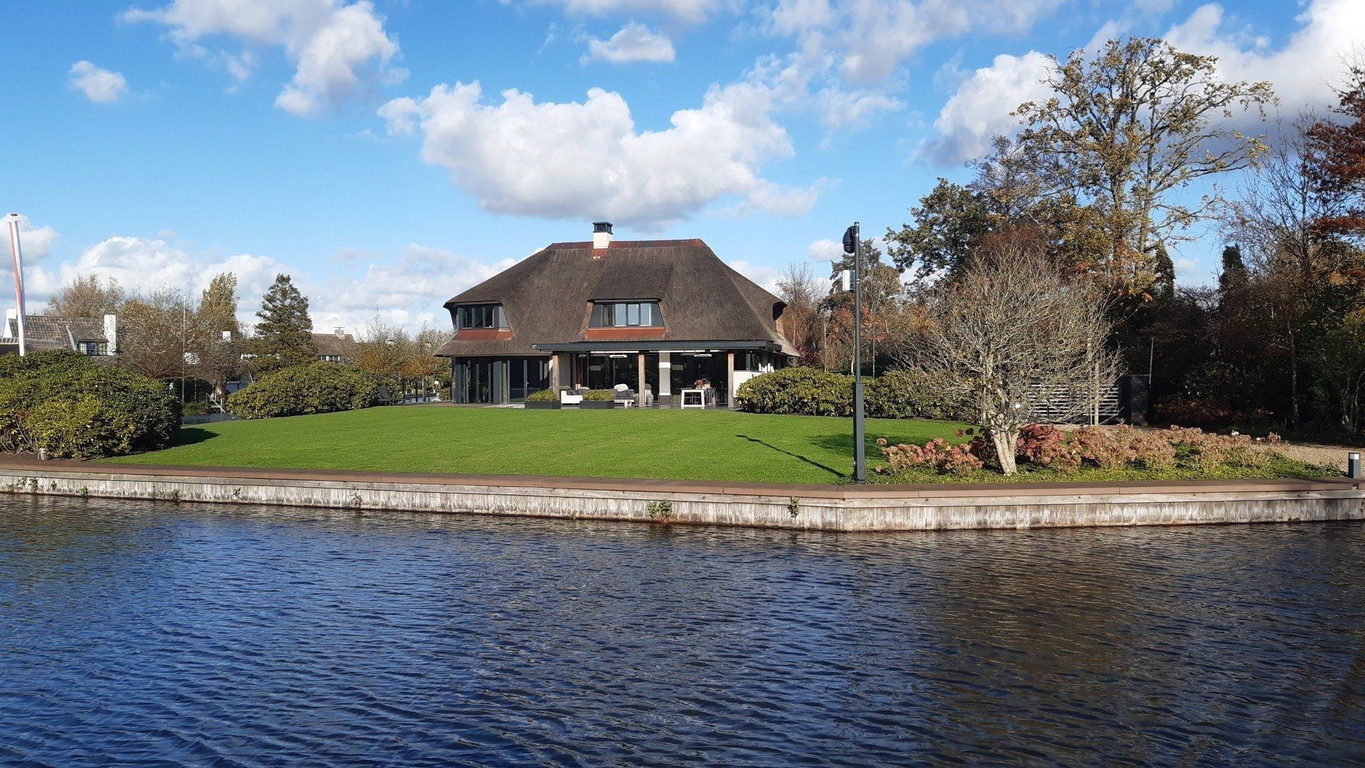 Huis met rieten dak op een grashelling naast een waterpartij, onder een blauwe hemel met wolken.