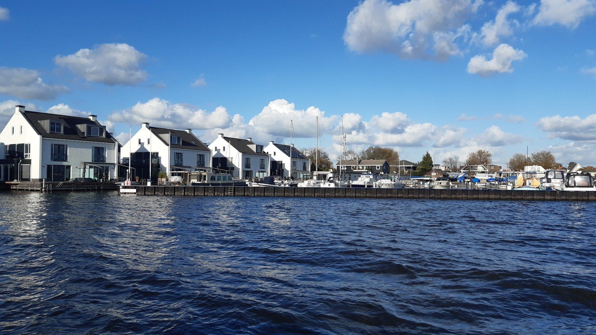 Een rij witte huizen met steigers en boten langs een blauw wateroppervlak, onder een gedeeltelijk bewolkte hemel.