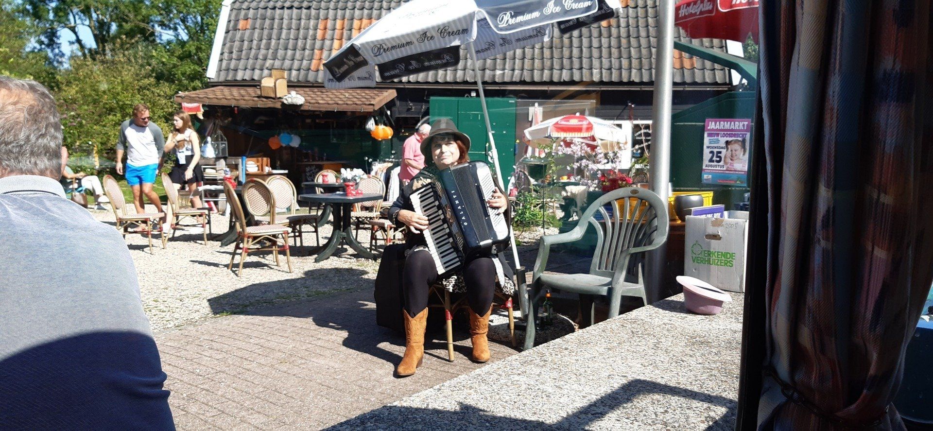 Iemand speelt accordeon onder een paraplu, in de buitenlucht. Op de achtergrond zijn andere mensen en een gebouw te zien.