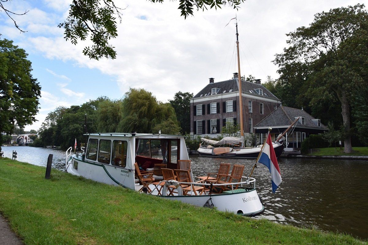 Boot op een rivier voor een grote buitenplaats met een Nederlandse vlag aan de zijkant.