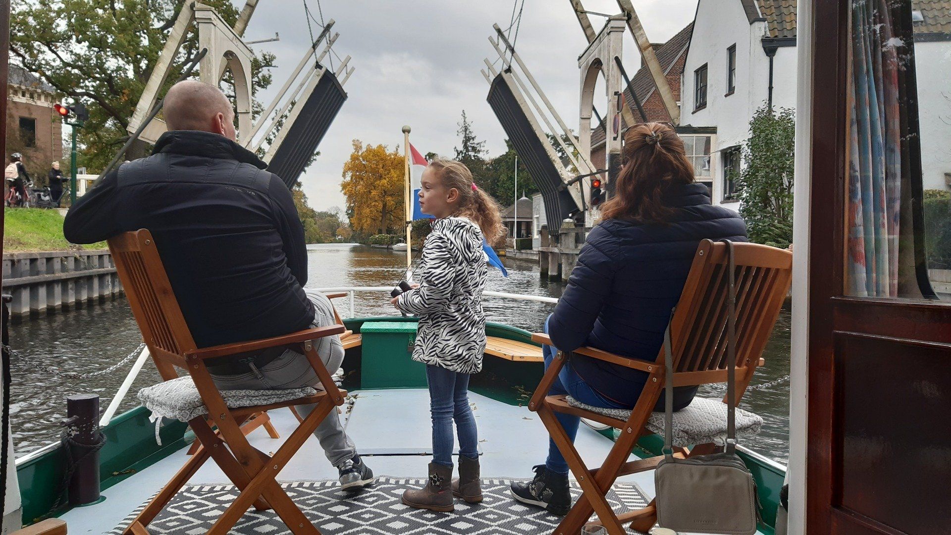 Een gezin in een boot vaart langs een open brug over een rivier. Het is een bewolkte dag.