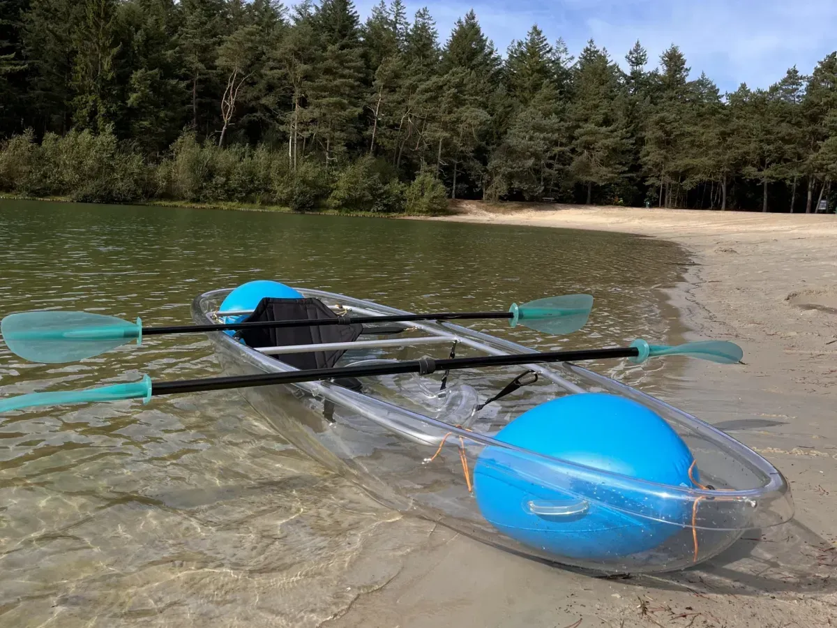 Een doorzichtige plastic kajak met twee felblauwe drijvers ligt op een zandstrand aan een meer met bomen op de achtergrond.