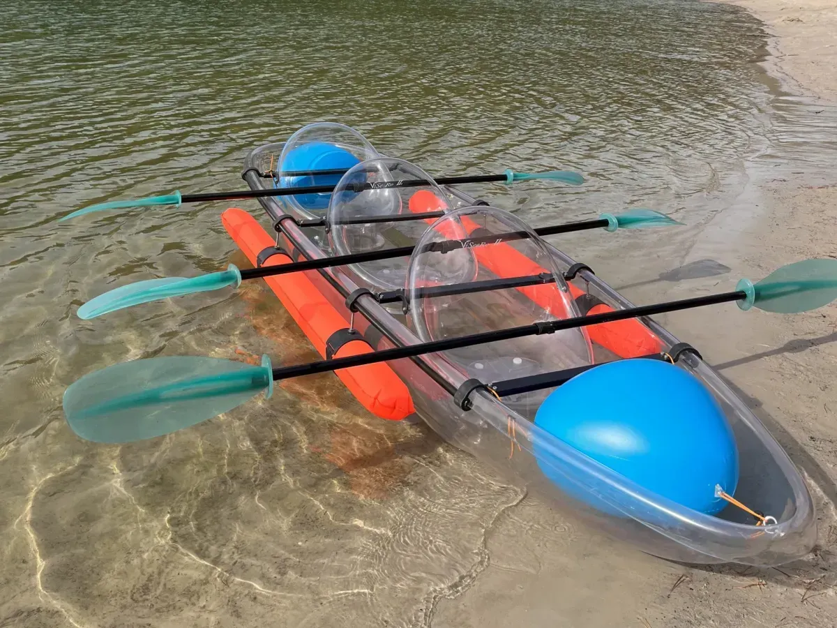 Een transparante tweepersoonskajak met felblauwe en oranje stootkussens, die op een zandstrand aan het water ligt.