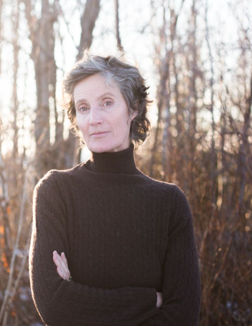 Woman in brown turtleneck, arms crossed, outside with bare trees in background.
