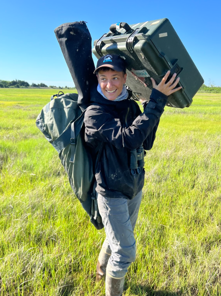 Person carrying gear across a grassy field under a bright sky.
