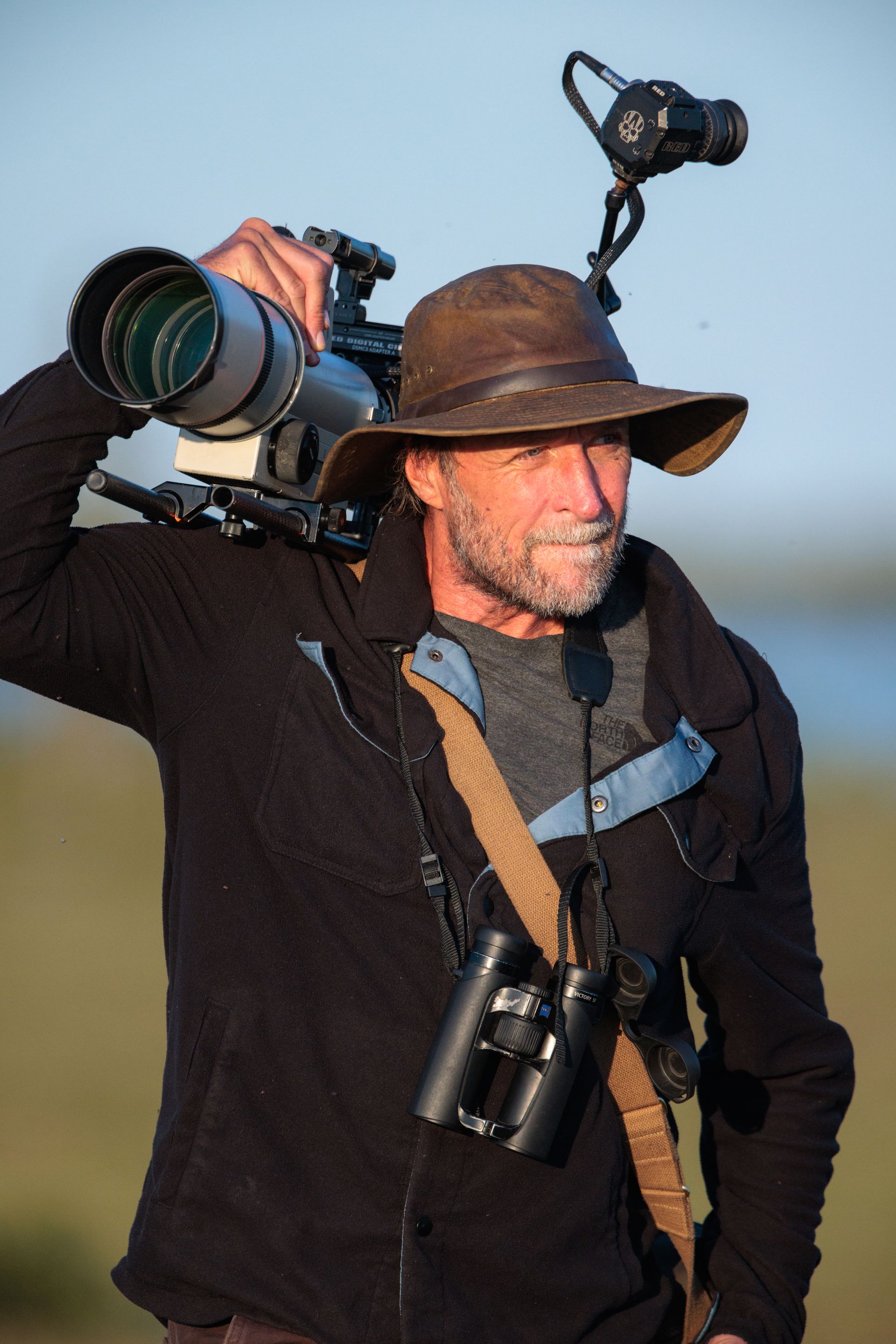 Man with camera and binoculars, wearing a hat, looking to the side outdoors.