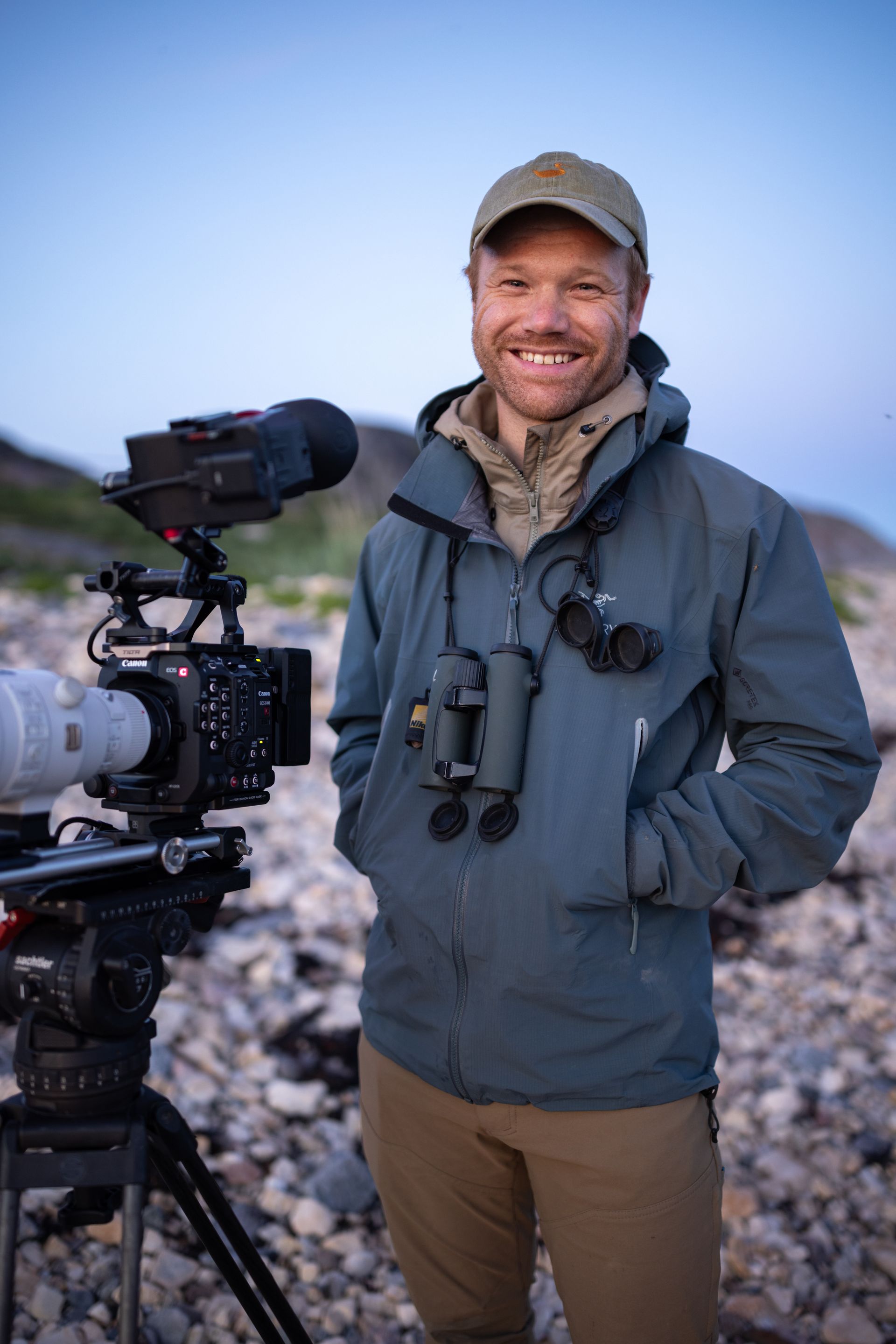 Man smiling, standing next to a camera on a tripod. Outdoors, wearing a cap, jacket, and binoculars.