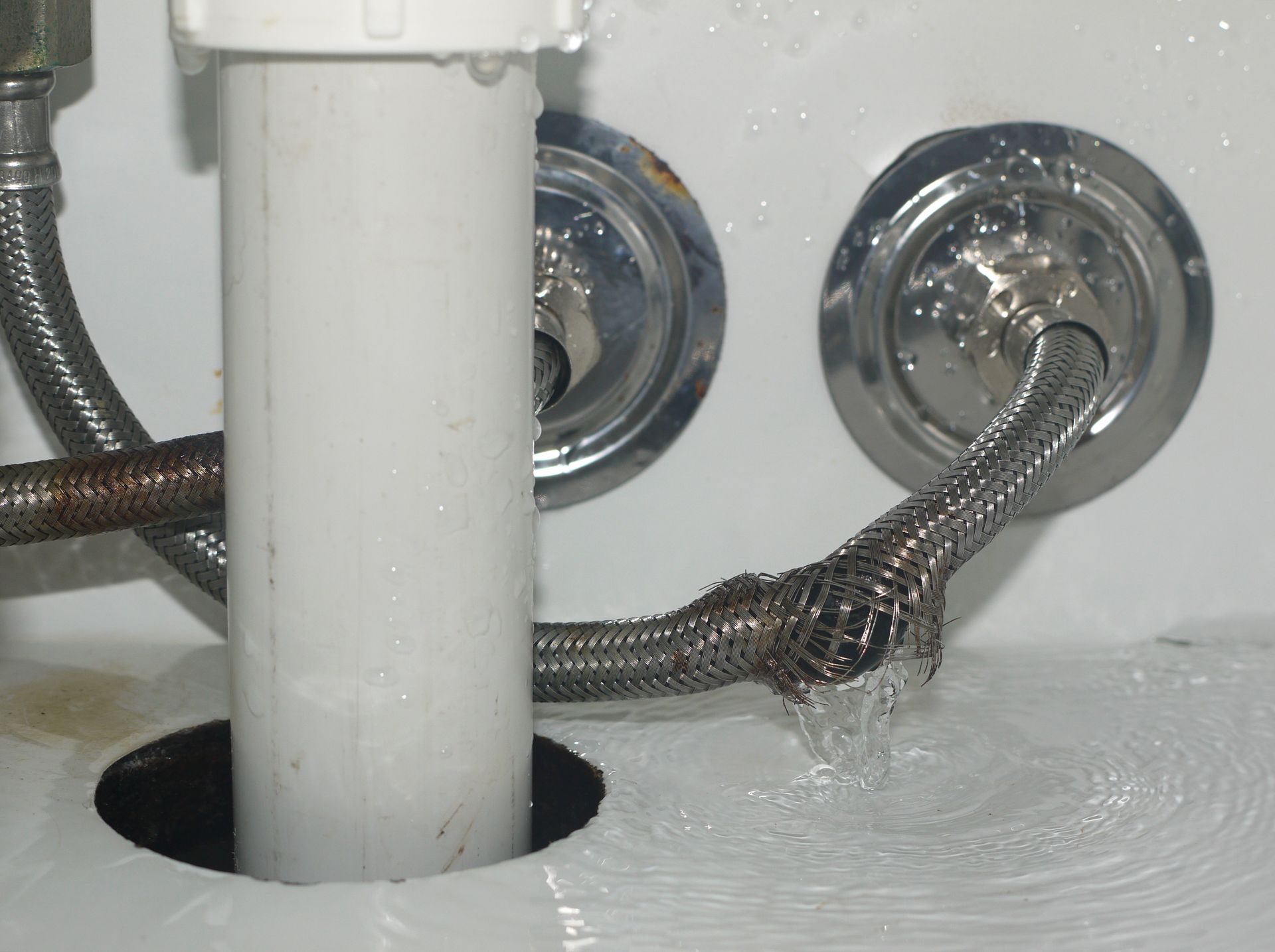 Under-sink plumbing with a braided hose leaking water around a white pipe and wall fittings
