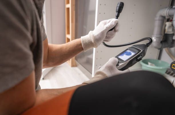 Person in white gloves uses a handheld meter to inspect a pipe under a sink cabinet.