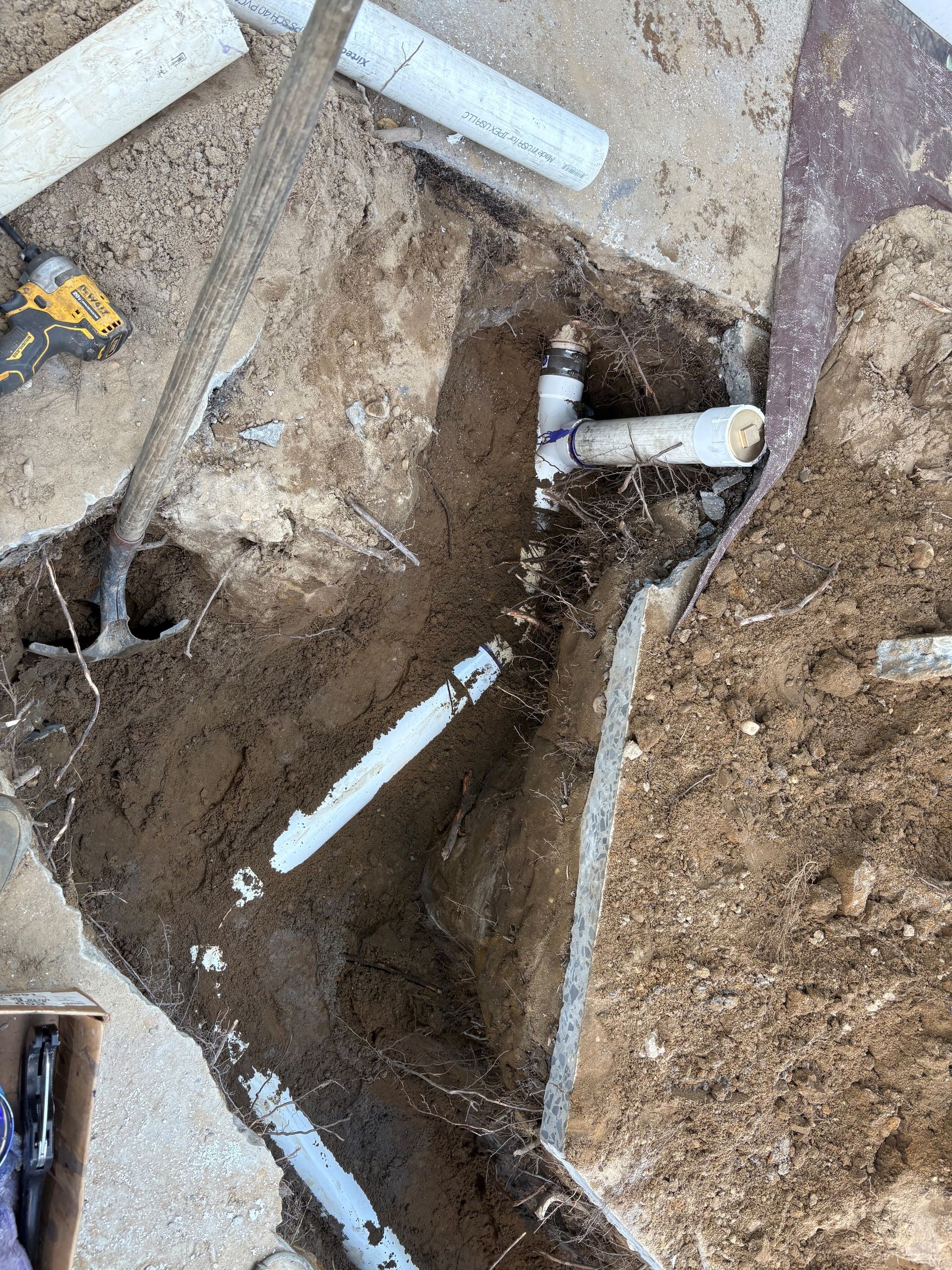 Trench excavation with white PVC pipes, dirt walls, and a yellow tool on the ground