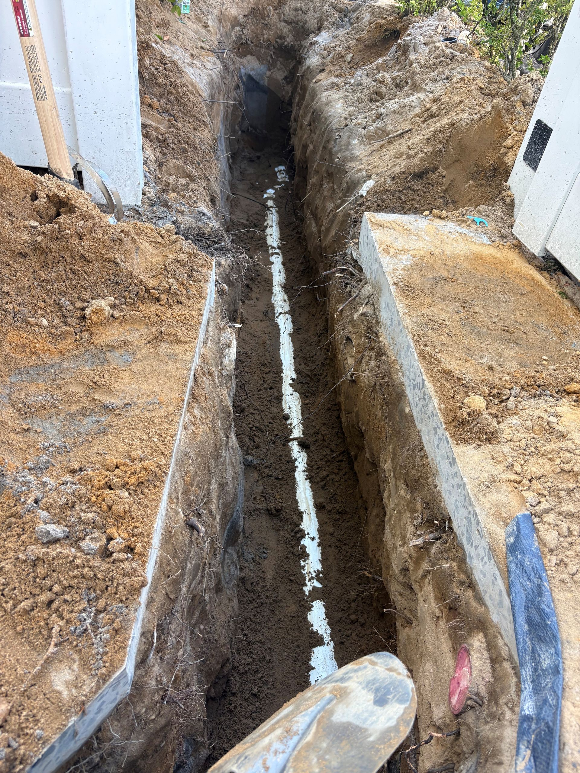 Narrow trench dug between two concrete surfaces, with exposed soil and a white cable inside.