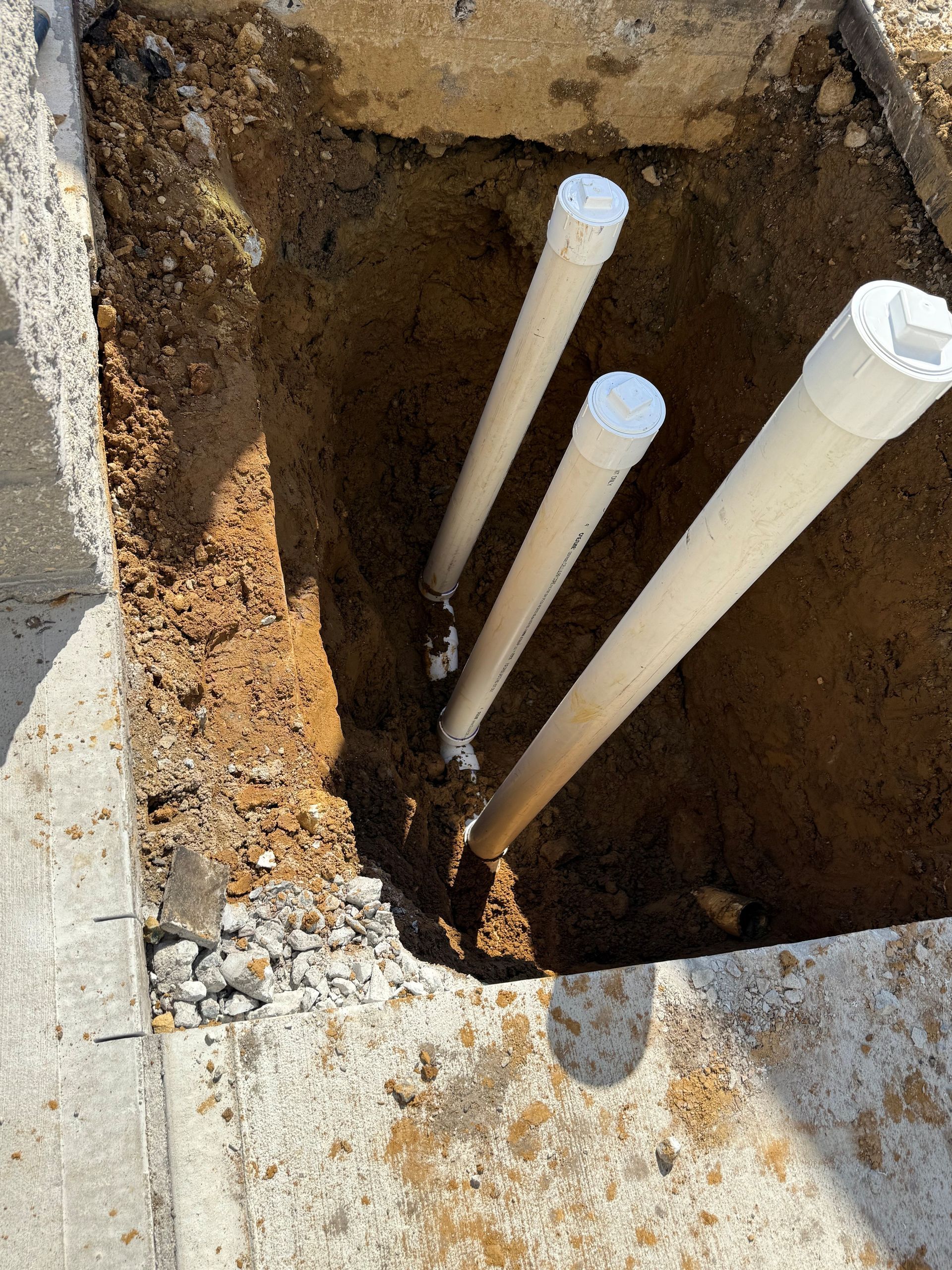 Open trench with four white PVC pipes rising from the soil beside concrete edges