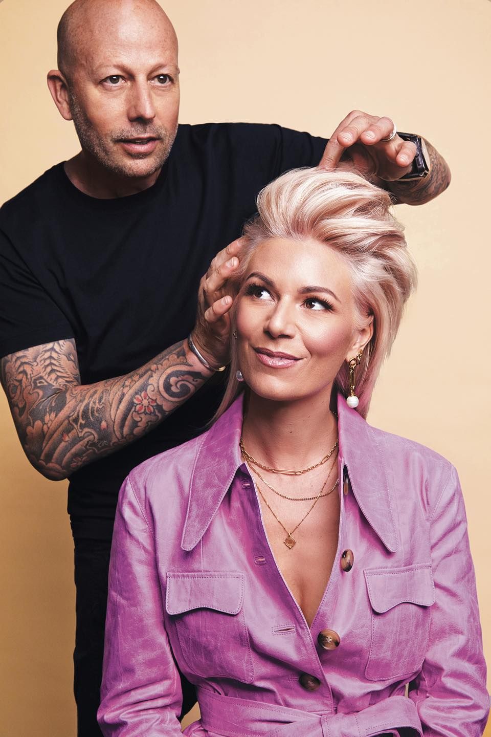 A stylist adjusting the hair of a woman with light pink hair; both are in front of a light yellow backdrop.