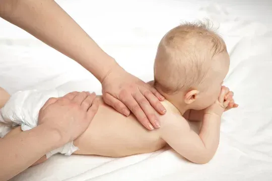 Mani che massaggiano la schiena di un neonato su un lenzuolo bianco. Il neonato è sdraiato prono, con lo sguardo rivolto verso il basso.