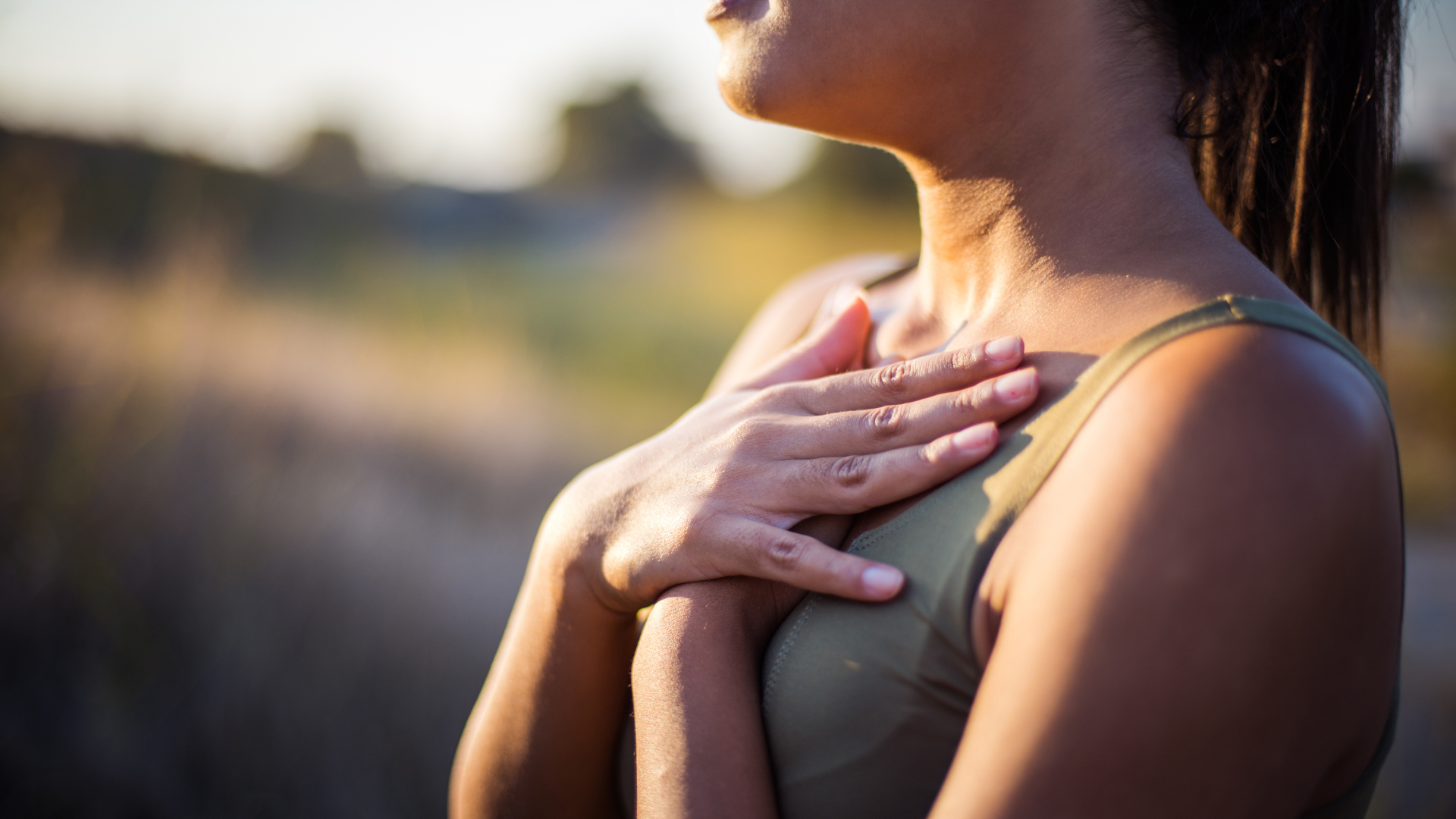 Woman outdoors with hand on chest, lit by sunlight.