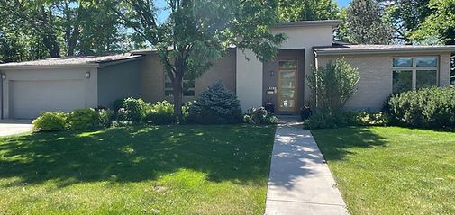 Single-story suburban house with a centered walkway, green lawn, and shrubs under a sunny sky