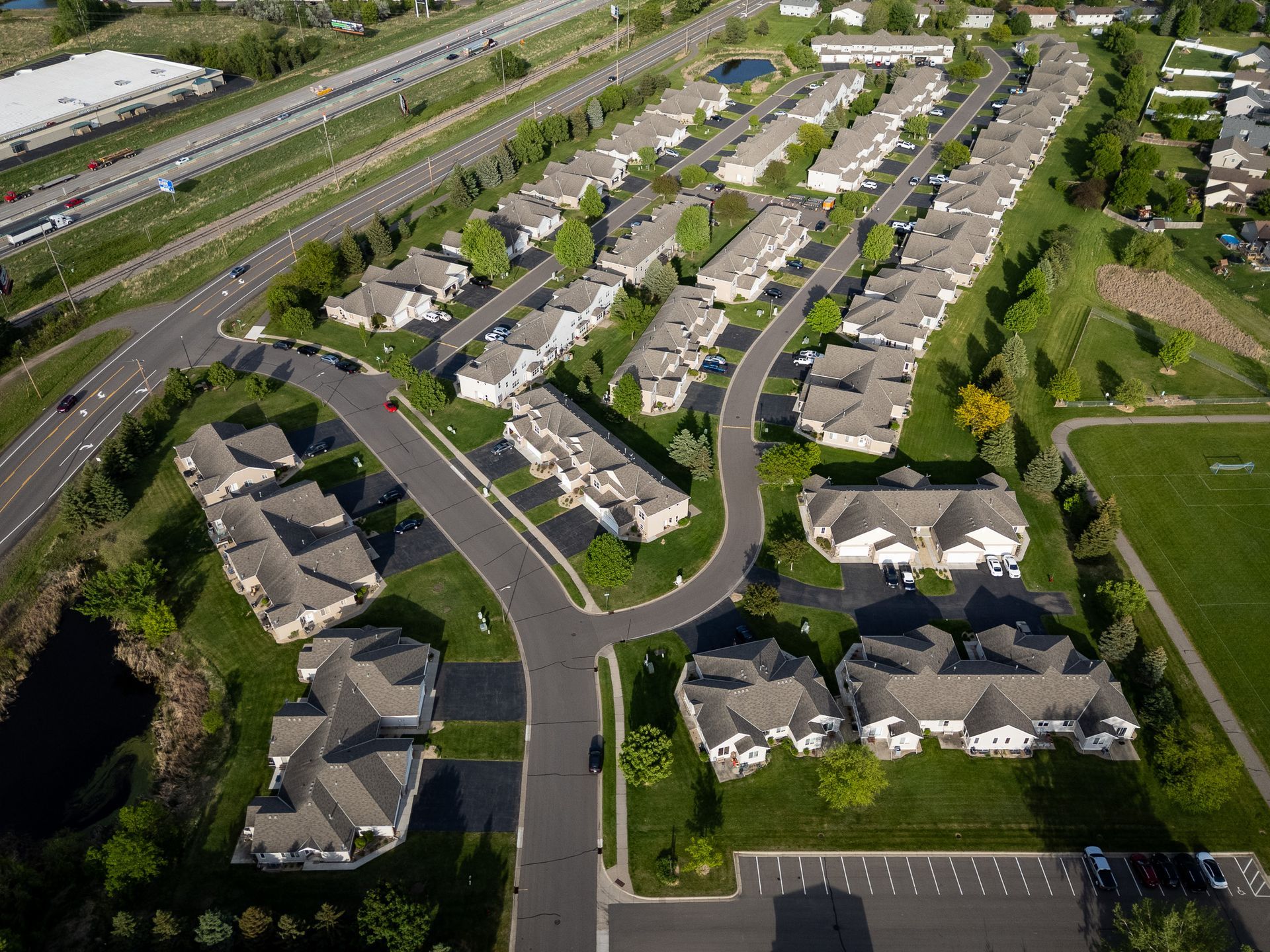 Aerial view of a suburban housing complex beside a highway, with curved streets and parked cars.