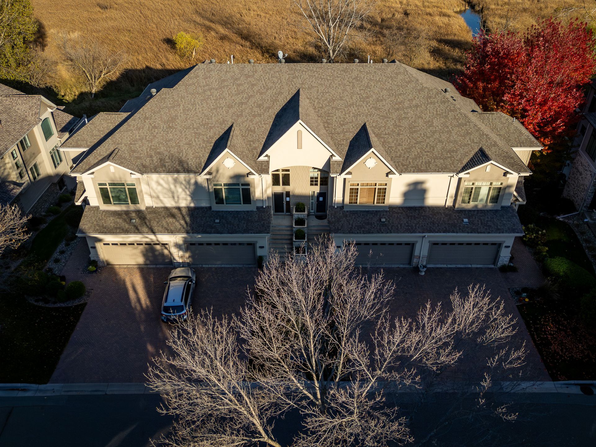 Aerial view of a large gray-roofed suburban townhouse with garages and a leafless tree in front