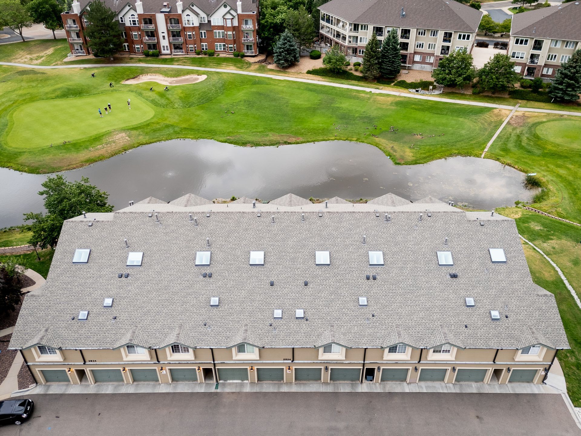 Aerial view of a clubhouse roof beside a pond and golf course, with houses in the background.