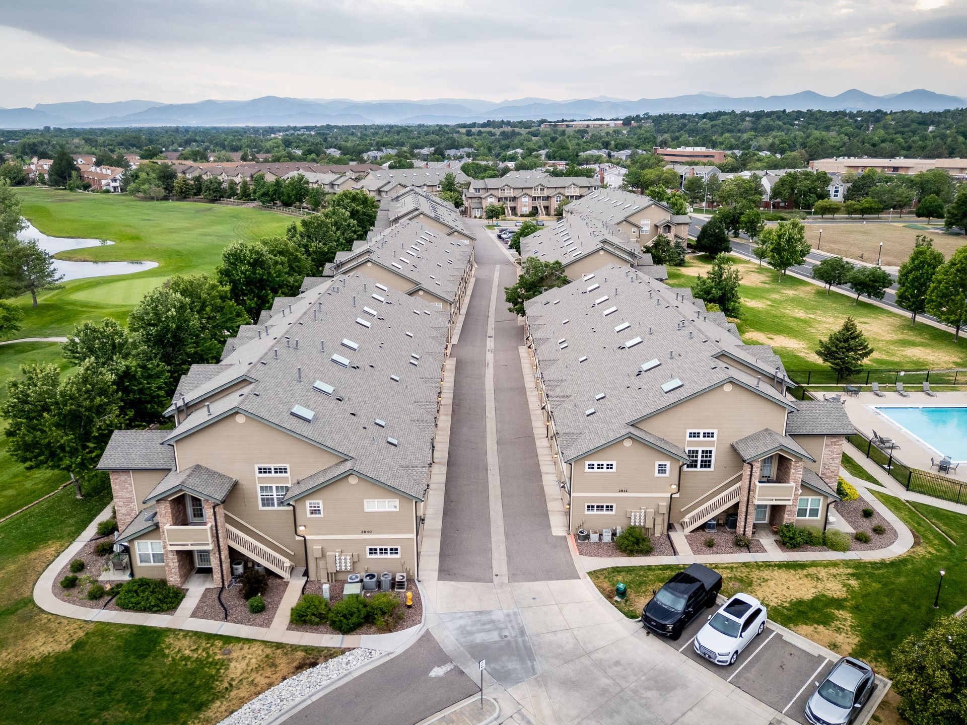 Aerial view of beige apartment buildings with a central driveway, parking, and a pool in a suburban neighborhood.