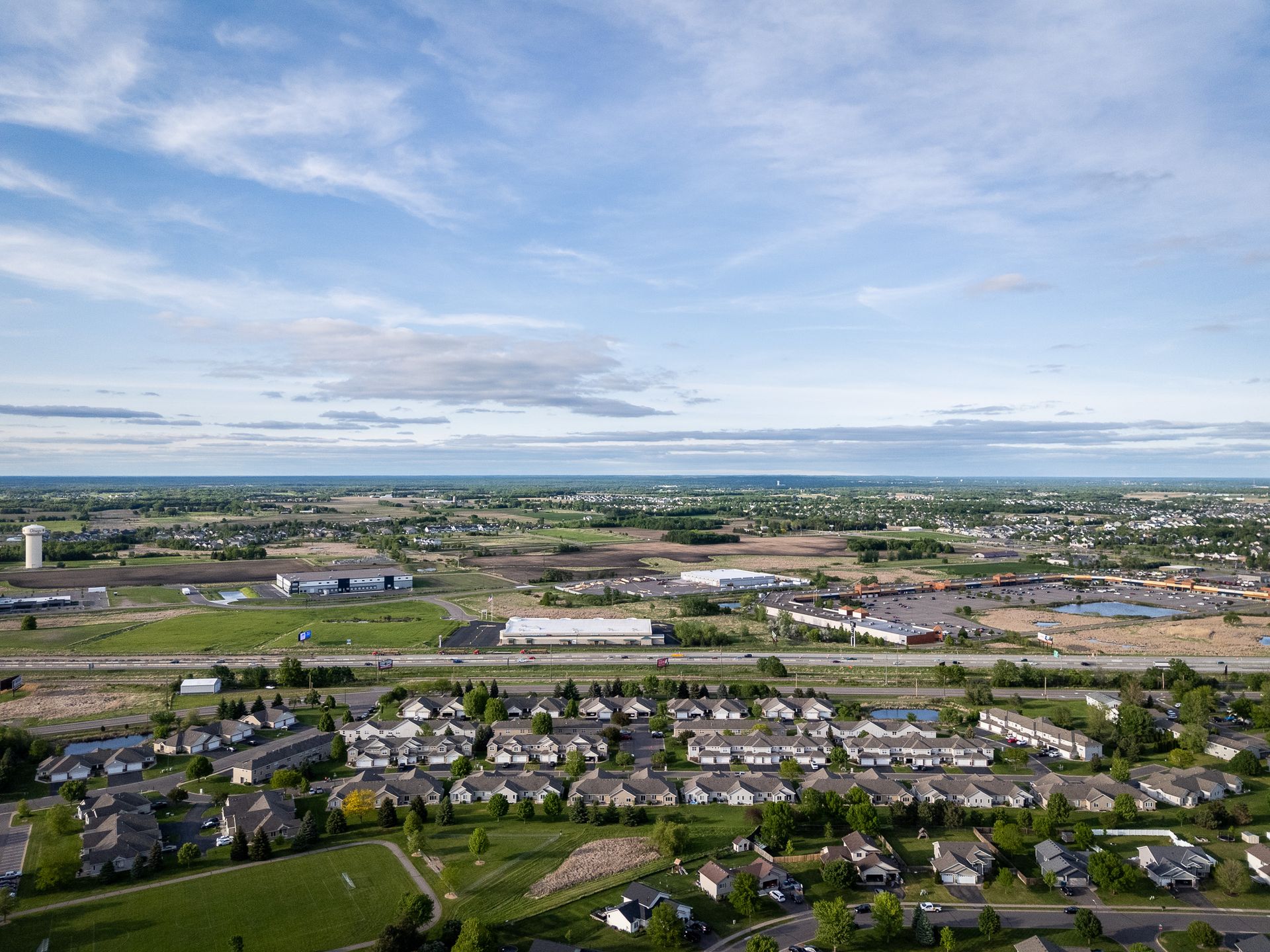 Aerial view of a suburban neighborhood with houses, roads, fields, and a distant coastline under a blue sky