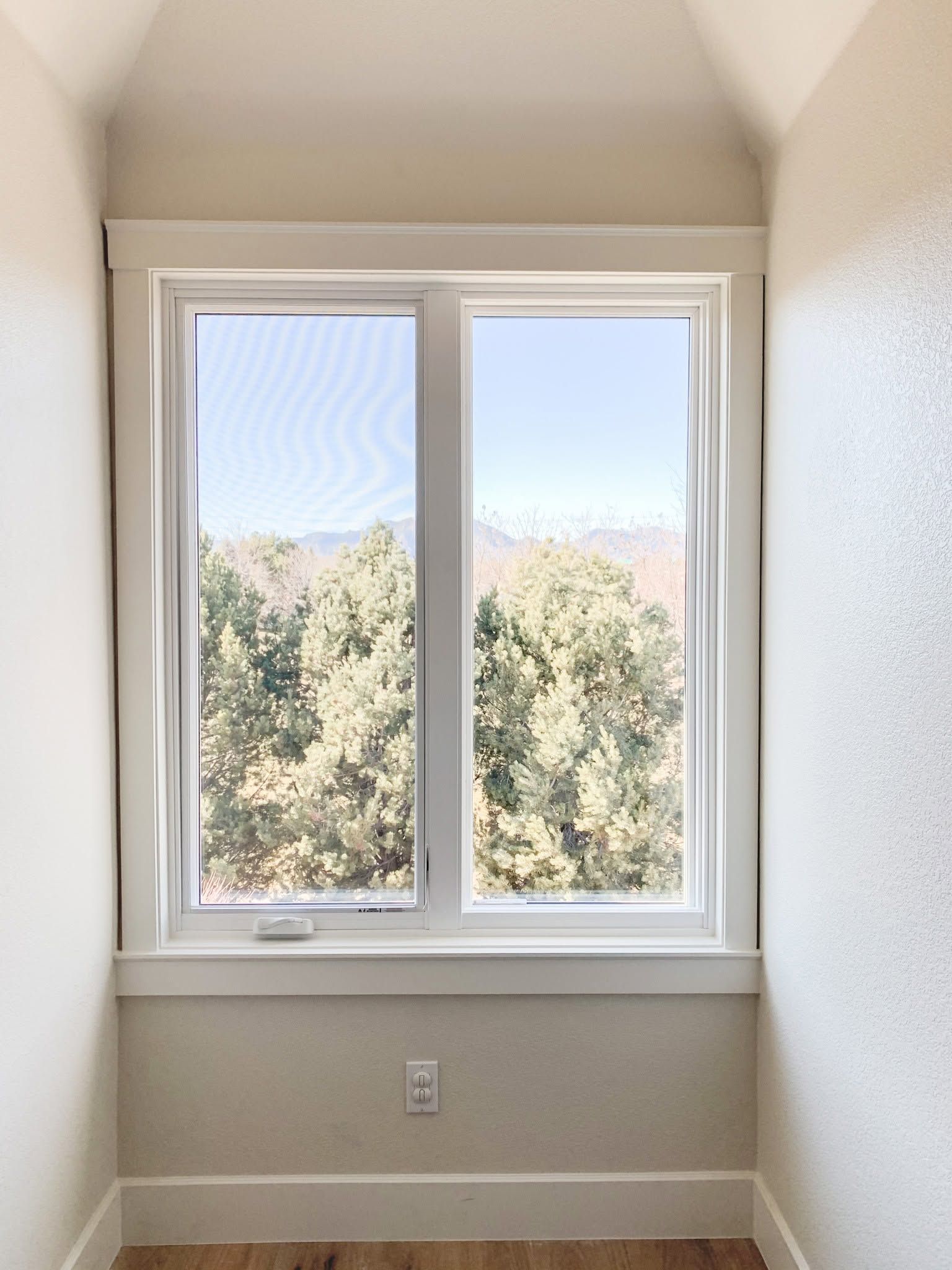 Small empty room with a white-framed window overlooking trees and a wall outlet below.