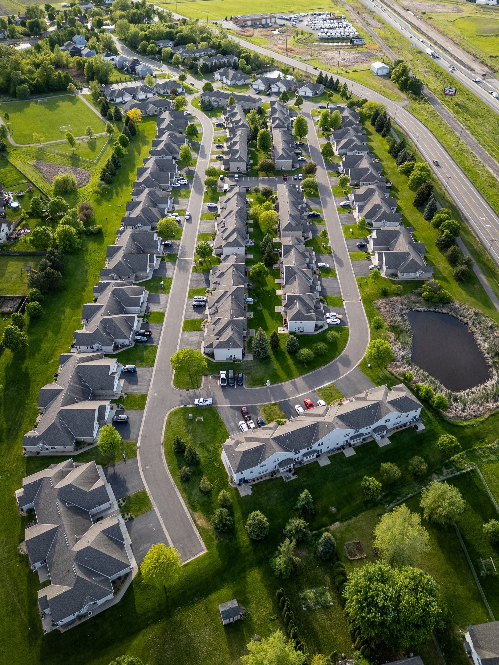 Aerial view of a suburban neighborhood with rows of houses, curving roads, trees, and a pond