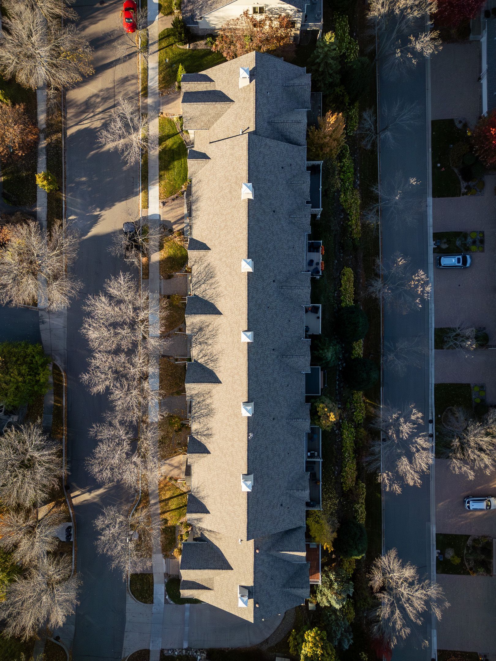 Aerial view of a long gray-roofed building bordered by trees, shadows, and a parking lot with a few cars.