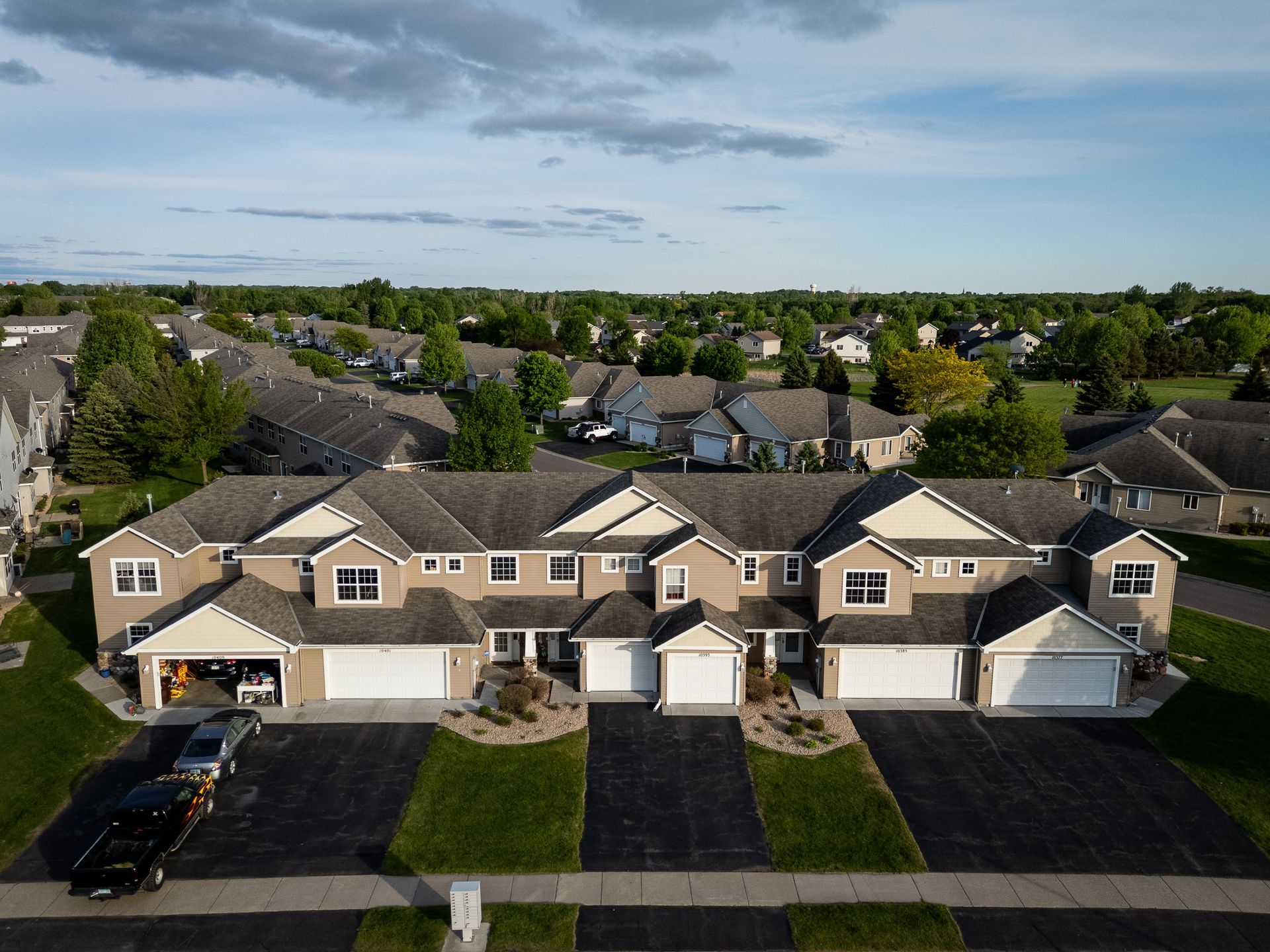 Aerial view of tan suburban townhouses with garages, driveways, and a tree-lined neighborhood under a blue sky